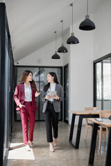 Two young businesswomen are walking through a modern office space, engrossed in a discussion while...