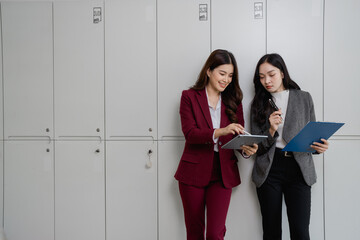 Two asian businesswomen are standing next to lockers in an office, smiling and discussing work while looking at a tablet and some documents
