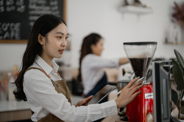 Young barista wearing apron using professional coffee grinder and holding tablet, managing orders in modern cafe with colleague working in background