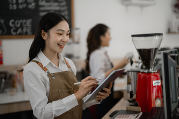 Young asian barista smiling and using digital tablet for managing orders and checking online sales in a modern cafe, with colleague working in the background