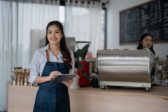 Smiling asian female barista wearing apron using digital tablet managing orders while standing at cafe counter, coffee machine and colleague working in background - Powered by Adobe