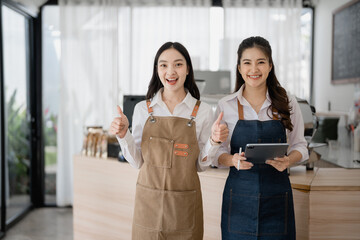 Two smiling Asian waitresses, dressed in bright aprons, giving thumbs up while working in their cheerful coffee shop, with one holding a tablet, radiating positivity and teamwork
