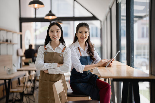 Two young smiling waitresses are posing in their coffee shop, one is standing with arms crossed, the other is sitting on a chair at a table using a tablet - Powered by Adobe