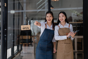 Two cheerful Asian female entrepreneurs, one pointing at the open sign and the other holding a digital tablet, celebrating the launch of their new cafe