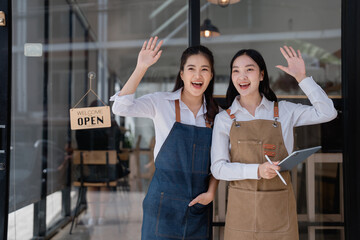 Two young smiling asian waitresses wearing aprons are waving at the entrance of their coffee shop or restaurant, in front of a welcome open sign, one of them is holding a tablet with a pen