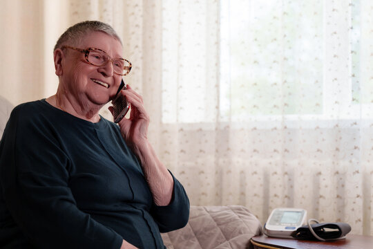 An elderly retired woman with gray hair, short haircut, wearing glasses, smiling, talking on the phone discussing health, next to a device for measuring blood pressure.