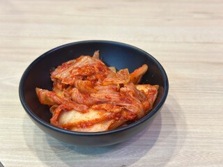 Authentic Korean Horizontal Shot: Spicy Fermented Napa Cabbage Kimchi (Baechu Kimchi) Served as a Side Dish in a Matte Black Bowl on a Light Wooden Table.