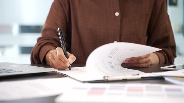 Close up of female hand signing documents at a desk at a workplace in a business office. Confident businesswoman manager looks through the folder with documentation and signs the contract with a pen
