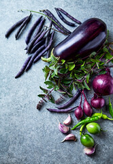 Different purple and green vegetables and herbs on gray stone background. Healthy organic food ingredients. Copy space