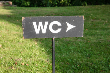  Black WC sign with white letters and arrow pointing direction, mounted on a metal post in grassy outdoor setting. Public facilities and guidance