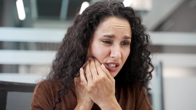 Businesswoman suffering from toothache sitting at workplace in modern business office. Upset female employee holds her hand near her cheek, massaging a sore spot. She needs help from dentist. Close up