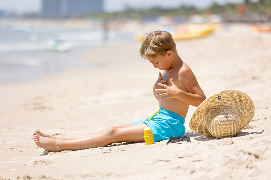 Mother and her son, cute blond boy, playing on the sea, hat, sunglasses, swimsuit, sun protection cream, sea and sand background. Concept: love, lifestyle, children, vacations