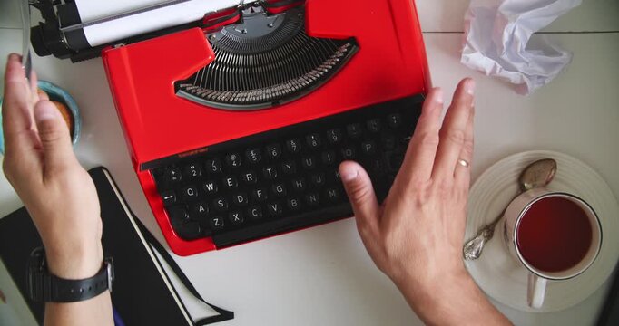 Hands typing on vintage red typewriter at desk with tea, cookies, notebook and crumpled paper. Concept of writing, creativity and retro style.
