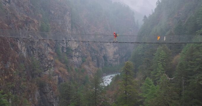 Drone view of Hillary Bridge near Monjo on Everest Base Camp trek, Nepal. A tall suspension bridge in rain shows tourists crossing, looking down at river and Himalayan landscape with awe and thrill