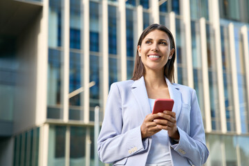 Young latin hispanic business woman using smartphone cellphone financial trading, looking dreaming aside. Indian middle eastern female entrepreneur businesswoman holding mobile cell phone outdoors. 