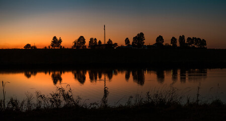 Fototapeta premium sunset over the horizon with the reflection of trees and a tower in the calm water of the lake