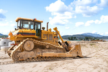 Heavy yellow bulldozer at a construction site on a sunny day. Large tracked machine with blade and cabin, parked on sandy ground with pipes and mountains in the distance. Industrial concept of earthmo