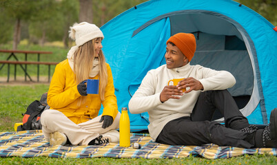 Interracial couple relaxing and drinking hot beverages outside their tent during camping trip
