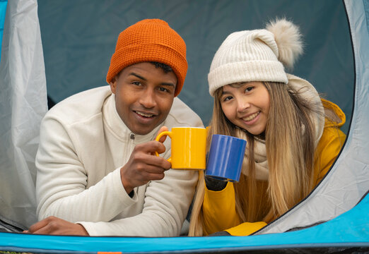 Smiling couple wearing winter clothes toasting with mugs inside tent during camping trip - Powered by Adobe
