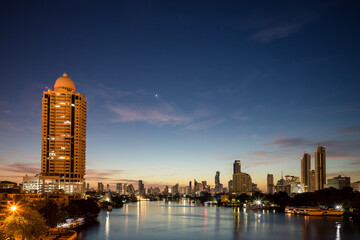 Twilight view of Bangkok skyline along the Chao Phraya River before dawn. Condominiums and hotels illuminated against the fading light, with city reflections shimmering in calm river.