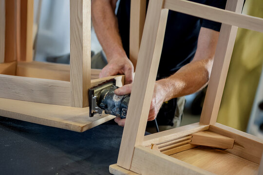 A craftsman sands the wooden frame of a chair with an orbital sander during the production process. The image shows woodworking and artisanal furniture.