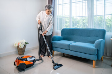 Senior Asian woman vacuuming living room floor with modern vacuum cleaner