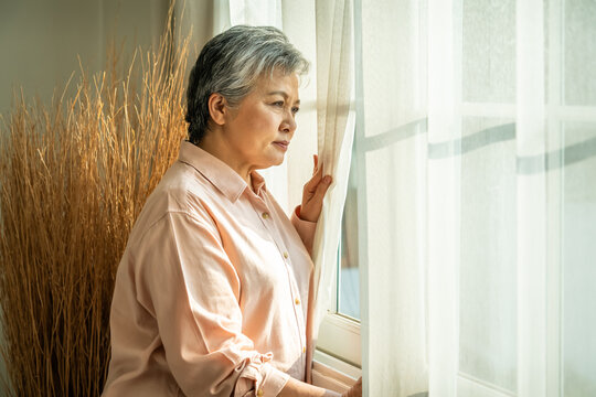 Senior elderly Asian woman looking out window with thoughtful expression in natural morning sun light