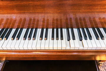 The row of black and white keys on piano keyboard reflect off the glossy brown wood of the musical instrument body. Vintage classical grand piano close-up.