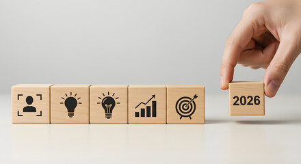 Hand placing a wooden block with 2026 next to business and idea icons symbolizing future planning and goals.