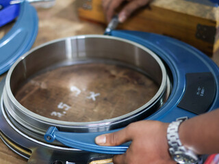 Close-up of a person's hands using a large Screw micrometer to measure a metal ring. The image captures the industrial process, focusing on precision, quality, and the tools involved