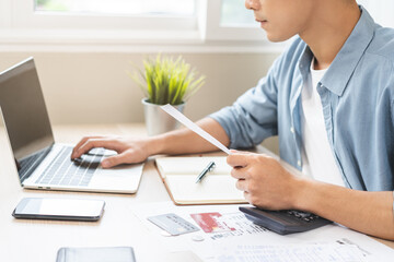 Stressed asian young businessman, employee male using laptop computer to calculate expenses, hand holding bills and receipt for to payment on table at home. Financial, finance of banking concept.