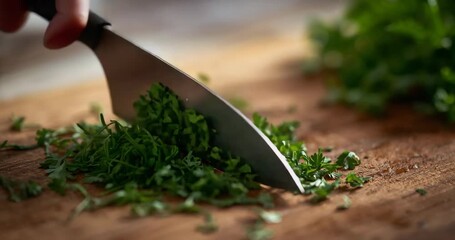 Cook gripping chef's knife mincing green herbs on wooden cutting board in kitchen for seasoning - Powered by Adobe