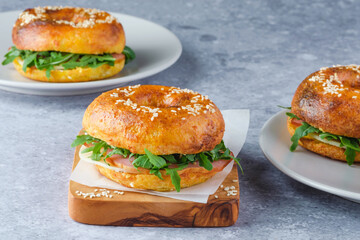 Three freshly baked bagels filled with greens and ham, on a wooden board and plates. Bagel breakfast sandwiches a zesty unique twist.