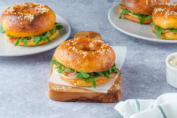 Three freshly baked bagels filled with greens and ham, on a wooden board and plates. Bagel breakfast sandwiches a zesty unique twist.
