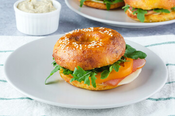 A delicious sesame bagel sandwich filled with ham, cheese, and fresh greens sits on a light plate....