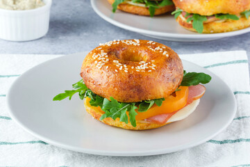 A delicious sesame bagel sandwich filled with ham, cheese, and fresh greens sits on a light plate. The meal is prepared on a textured surface, highlighting its freshness and color.