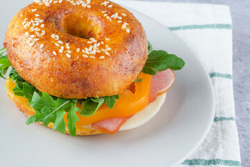 A delicious sesame bagel sandwich filled with ham, cheese, and fresh greens sits on a light plate. The meal is prepared on a textured surface, highlighting its freshness and color