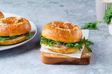 Freshly baked bagels filled with greens and ham, on a wooden board and plates. Bagel breakfast sandwiches a zesty unique twist. 