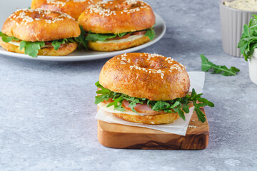 A delicious sesame bagel sandwich filled with ham, cheese, and fresh greens sits on a light plate. The meal is prepared on a textured surface, highlighting its freshness and color.