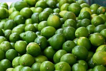 Fresh green limes in varying shades densely packed together, displaying round to oval shapes with natural surface texture and glossy reflections at fruit market.