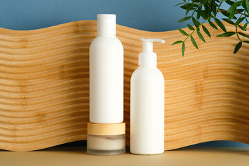 Minimalist skincare set with white bottles and wooden lid jar on wood-textured panel. Blue wall and green plant add natural contrast for clean beauty branding.