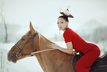 Elegant Asian woman in bright red dress sitting on a brown horse in snowy mountain landscape....