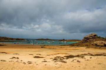Sandy beach and turquoise bay at low tide with anchored boats and dramatic pink granite rock formations in Brittany, France, under a cloudy summer sky.