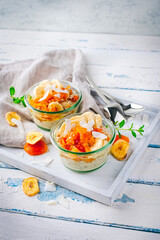 Overnight no-cooked oatmeal breakfast - oats soaked in almond milk with banana, coconut and apricot topping served as close-up in glasses on a wooden tray