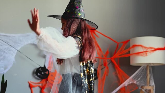 A girl in a white witch costume dances or casts a spell in a decorated room with Halloween garlands and fake cobwebs, looking down with a focused expression.
