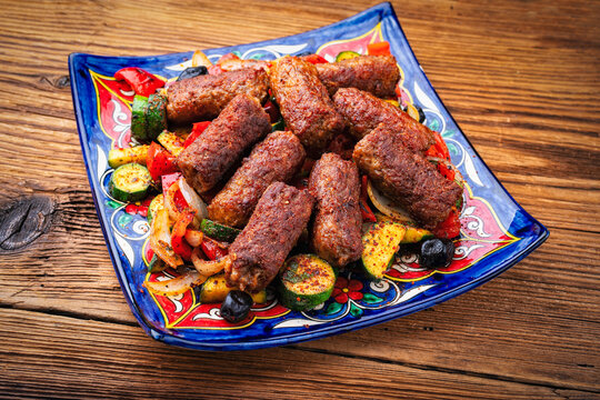 Traditional Croatian cevapi spicy meat ball rolls with troasted vegetables served as close-up on traditional rustic plate