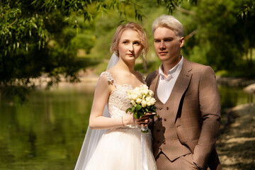 Couple in wedding attire poses by a serene lake surrounded by greenery