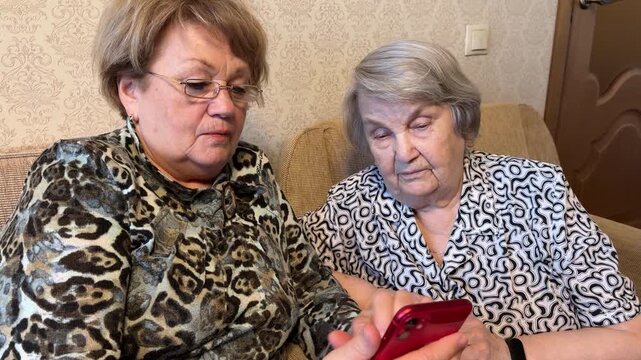 Two old women are watching photos on their phone while sitting on the couch at home