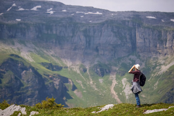 Hiker standing on alpine meadow in the Swiss Alps, looking at the dramatic mountain cliffs with a backpack and sun hat