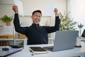 Excited businessman celebrating success at desk, raising papers in triumph while working on laptop.